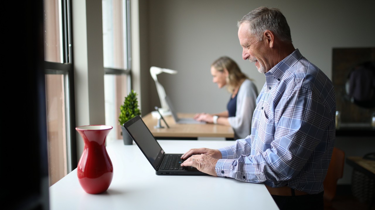 Man standing at desk with laptop, woman in background