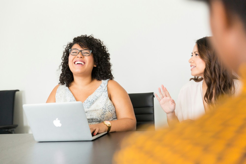 Two women in workplace meeting room smiling, with laptops in front of them
