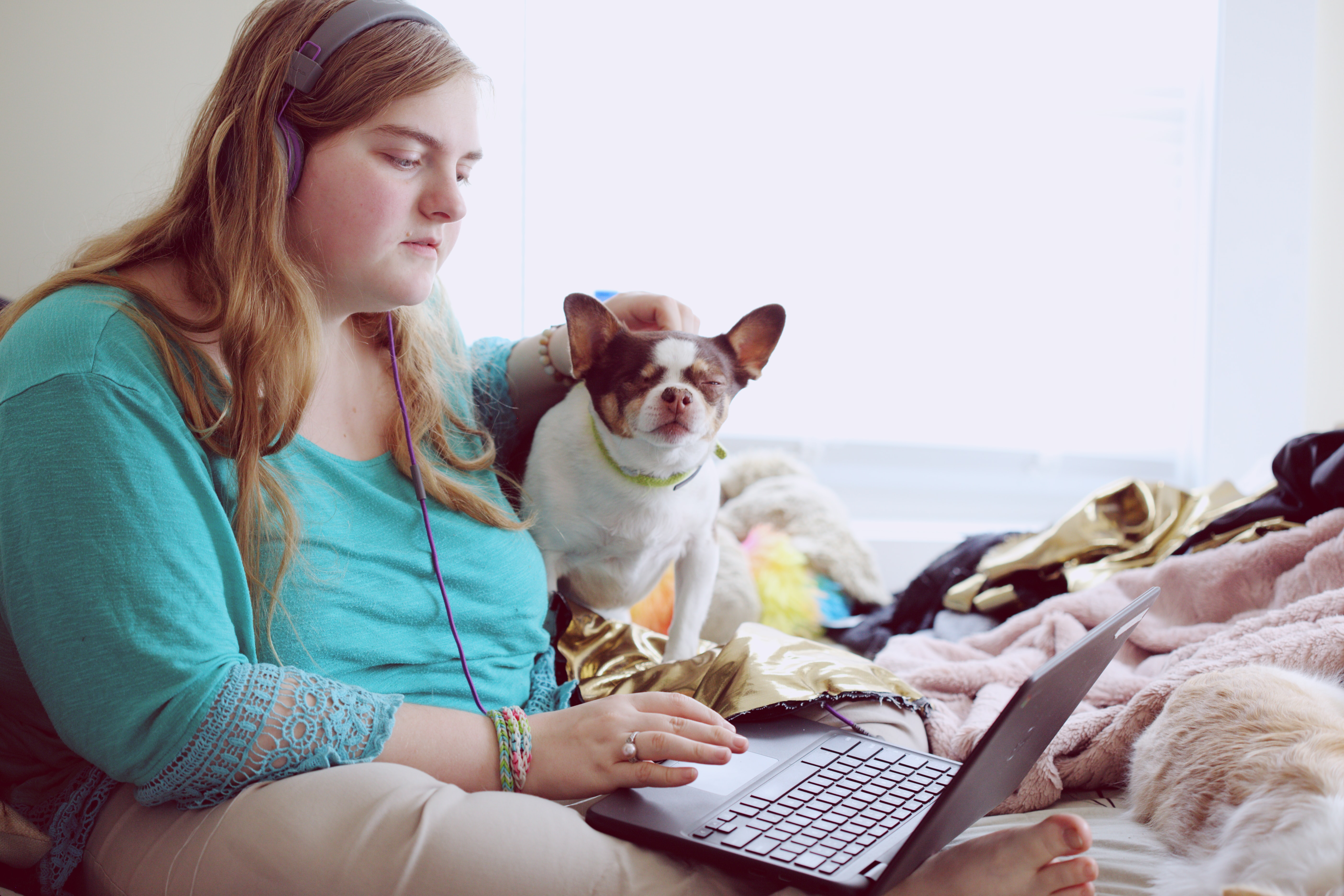 Image shows a 19 year old woman sitting on a bed with a laptop and a dog