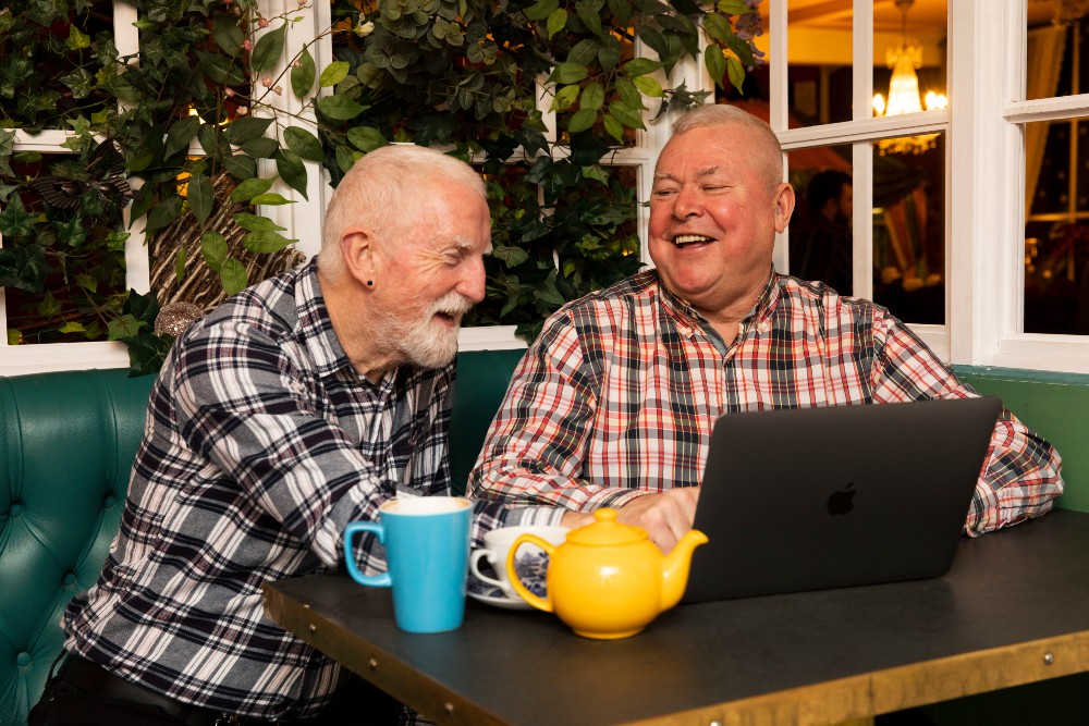 Two older men smiling in cafe setting looking at laptop on table
