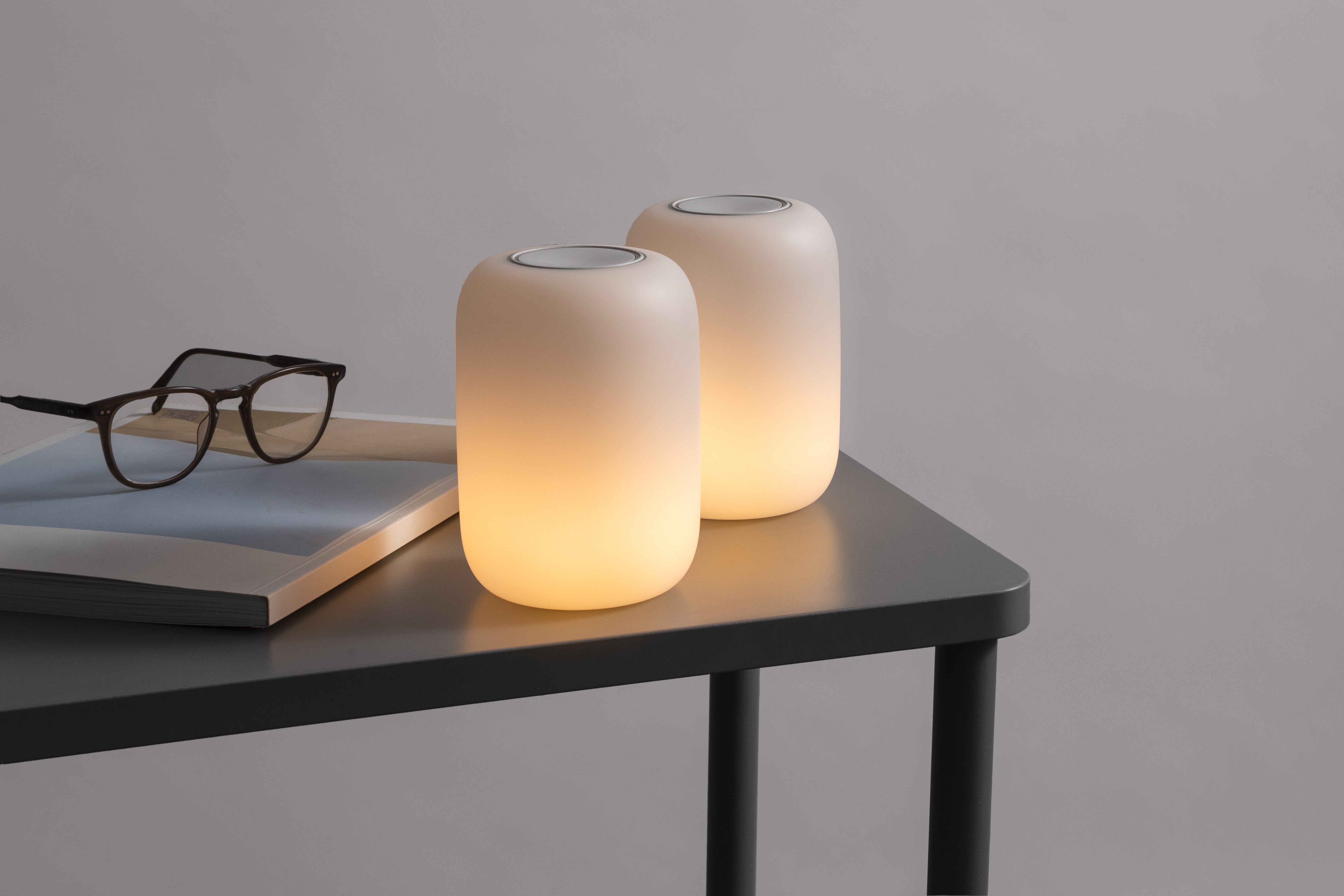 Two pill-shaped Casper Glow lights partially dimmed sitting on a bedside table with some books and a pair of glasses