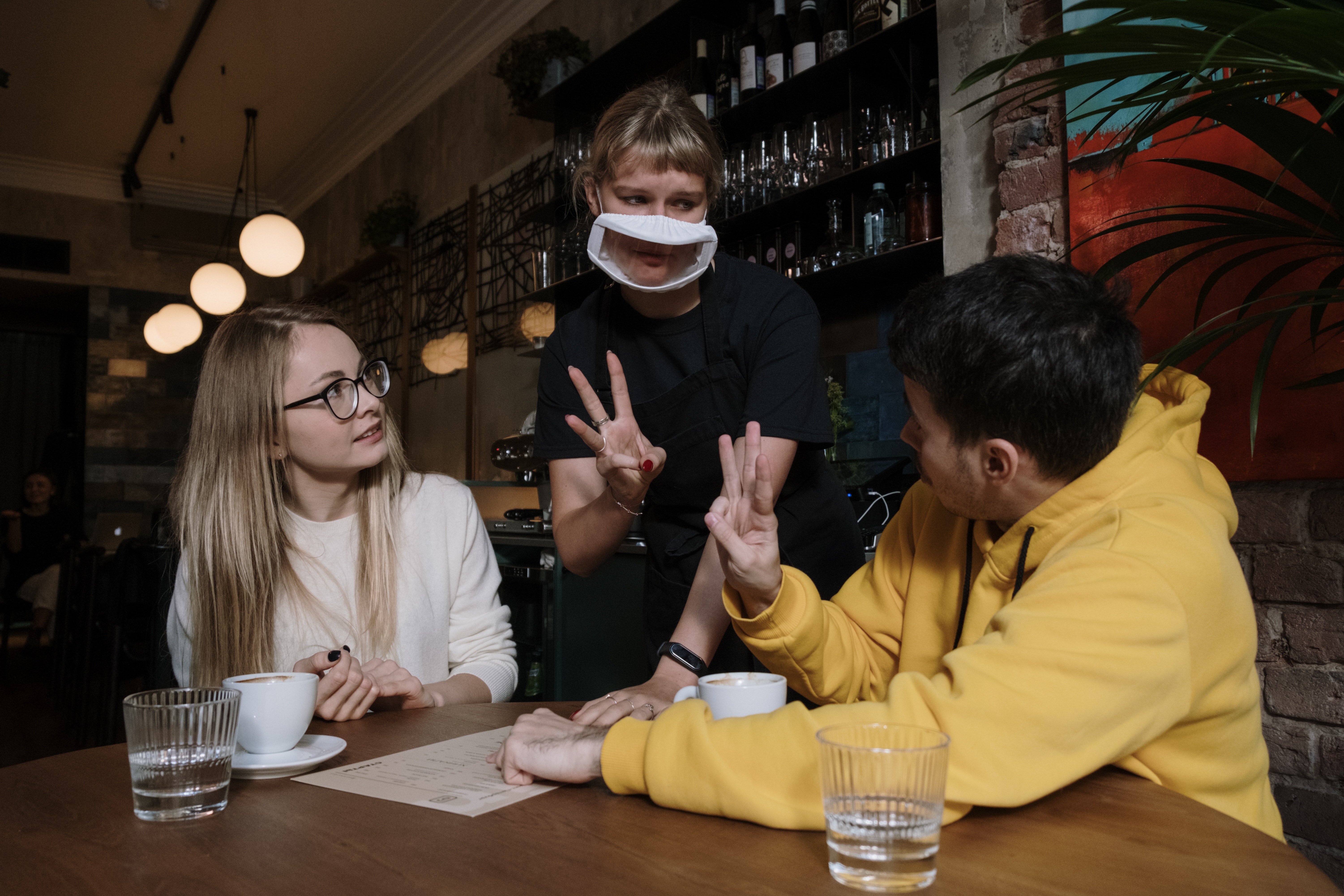 Image shows a woman in a coffee shop wearing a transparent mask signing to two customers at the tables