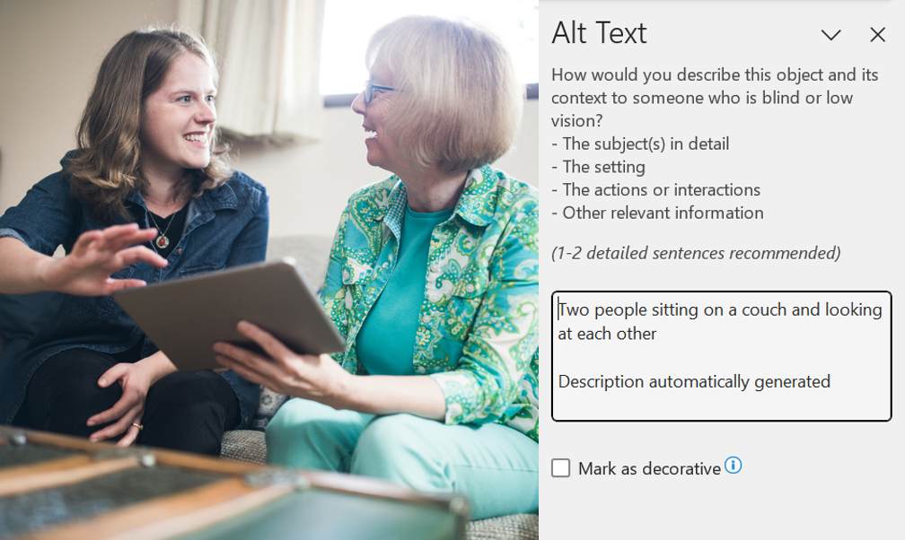 Screenshot of Microsoft word page with an image of a younger woman is helping an older woman navigate an iPad. They are sitting on a sofa and smiling at each other. The alt text editor is to the right, with the auto generated alt text reading 'Two people sitting on a couch and looking at each other'  