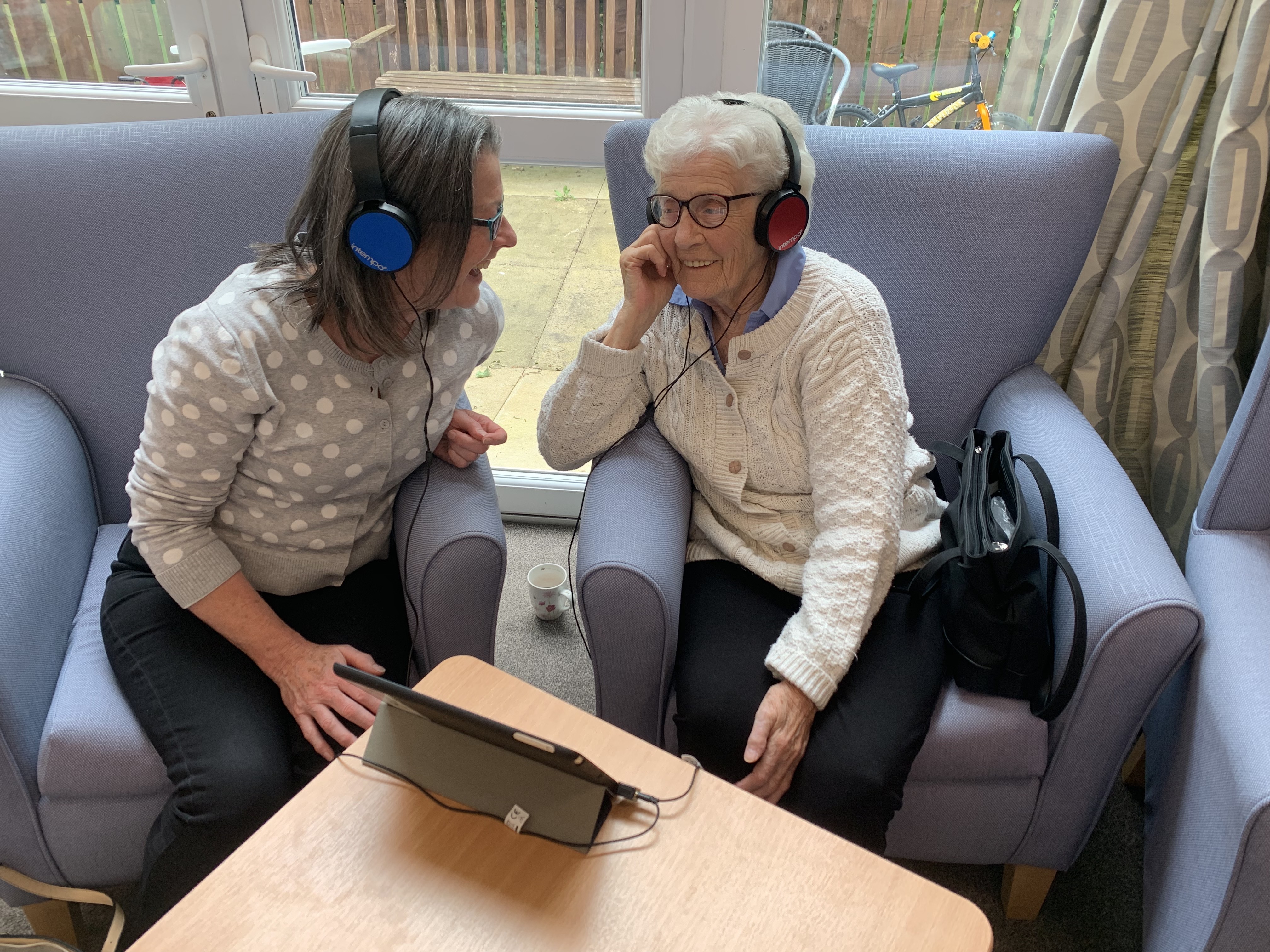 A young person and an older person both wearing headphone connected to a laptop. They are laughing.