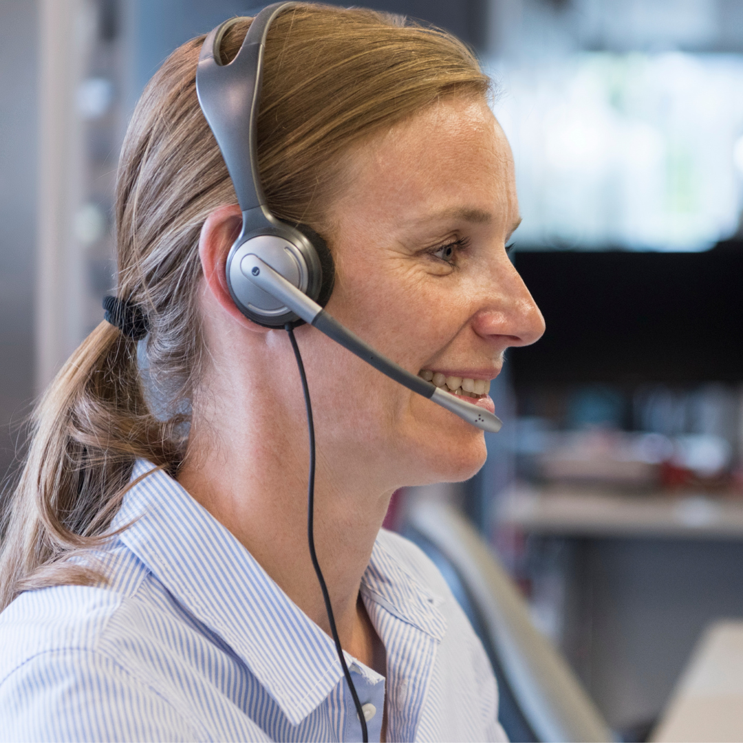 A person smiling at their desk whilst wearing a wired headset,