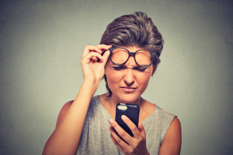 A woman lifting her glasses off her face, struggling to read something on her phone