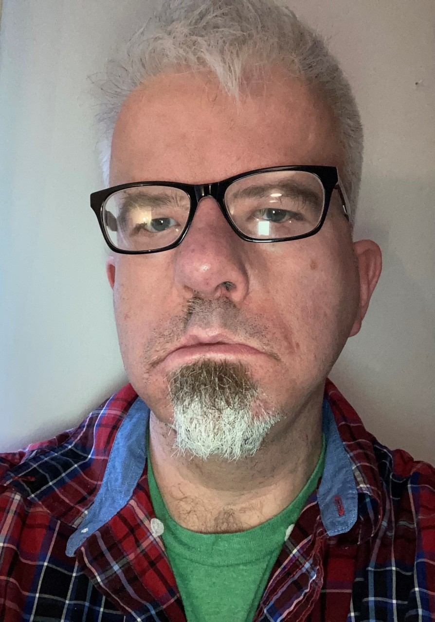 A greying 50 year old man in a checked shirt with a grey beard and black rimmed glasses