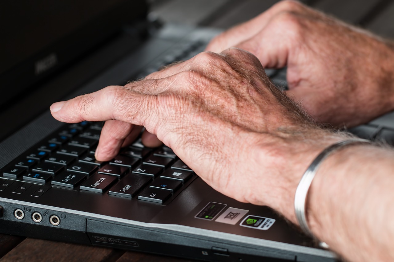 A picture of some older hands resting on a keyboard. There is a bracelet of the style worn for joint pain.