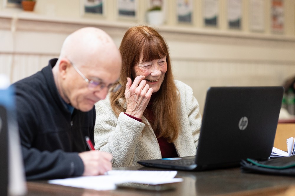 Older lady and man smiling and looking at laptop on desk in informal setting