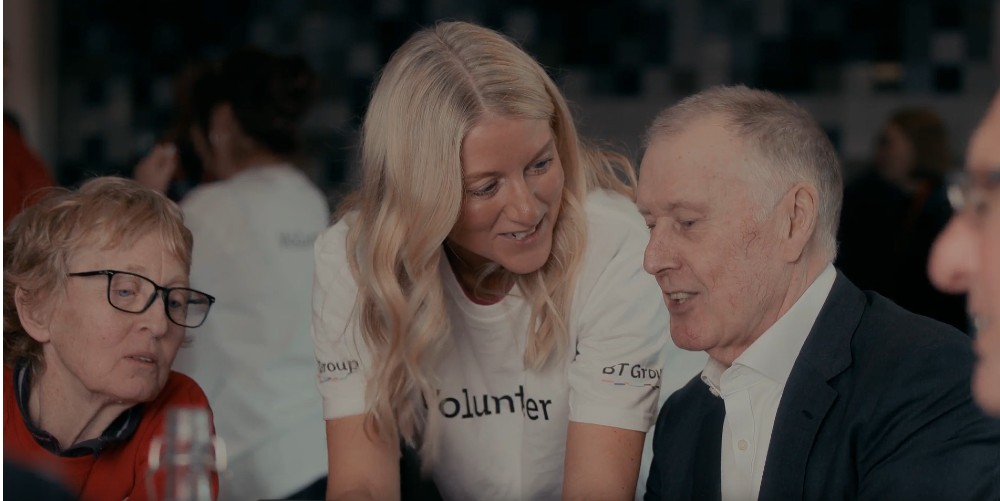 Sir Geoff Hurst looks at digital device while sitting at table with female volunteer and older woman