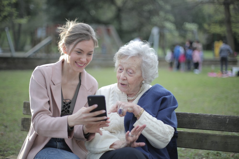 Younger woman shows older woman something on a phone, on a park bench