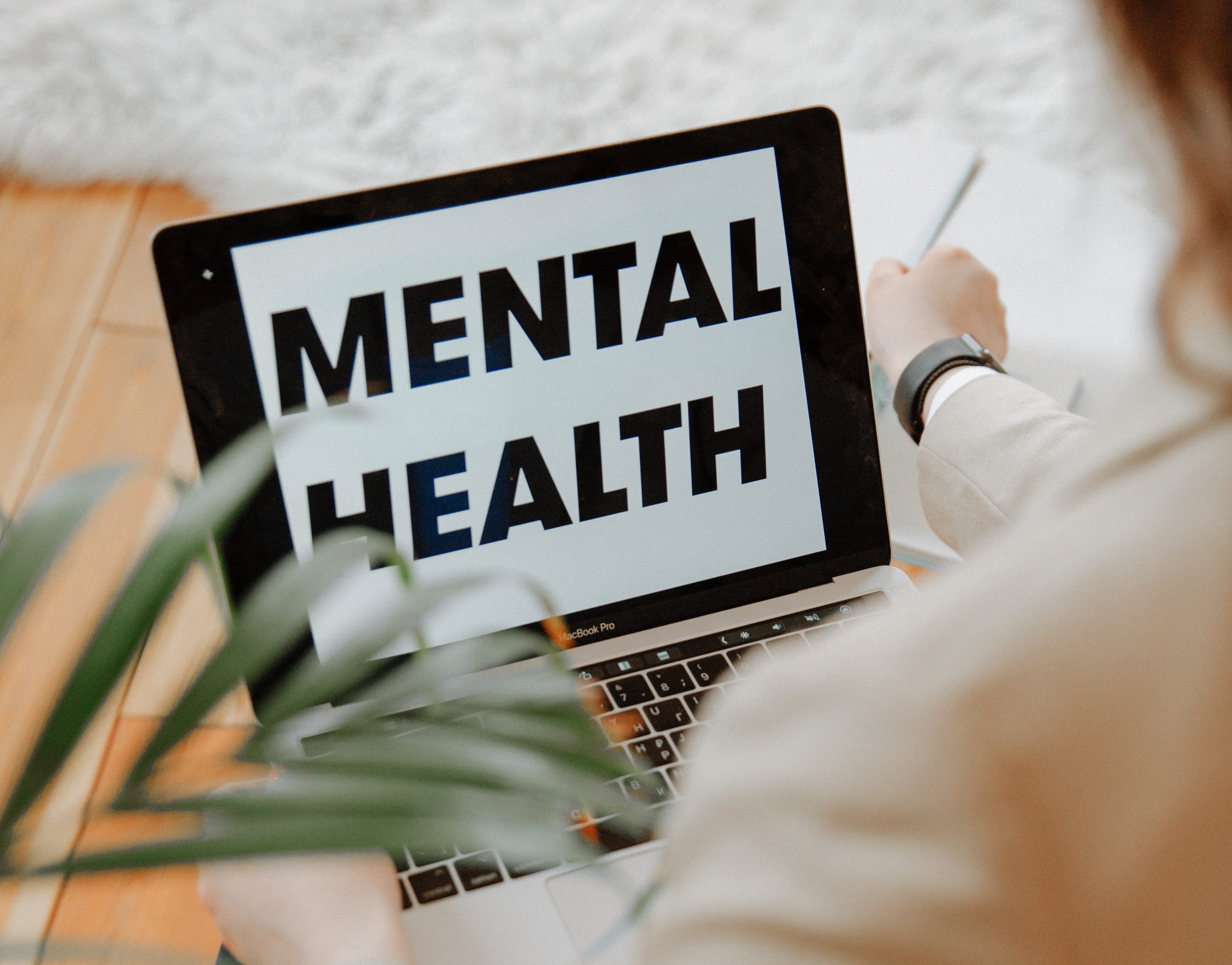 Image shows the top of a laptop with the words "Mental Health" written on it and viewed over the shoulder of the user who is out of focus.