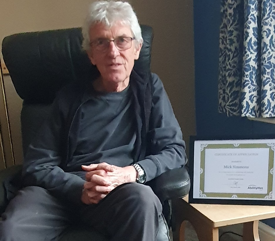 Mick Simmons sitting on armchair with desk showing framed certificate alongside him