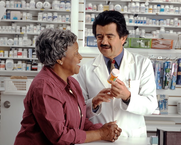 A picture of a pharmacist handing some pills to a woman. Rows of pills visible in the background.