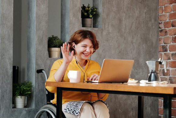 Woman in wheelchair smiling and waving while using a laptop