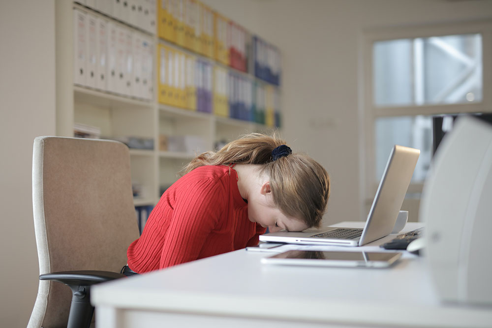 Person with head slumped on keyboard at desk in office environment, showing she is tired